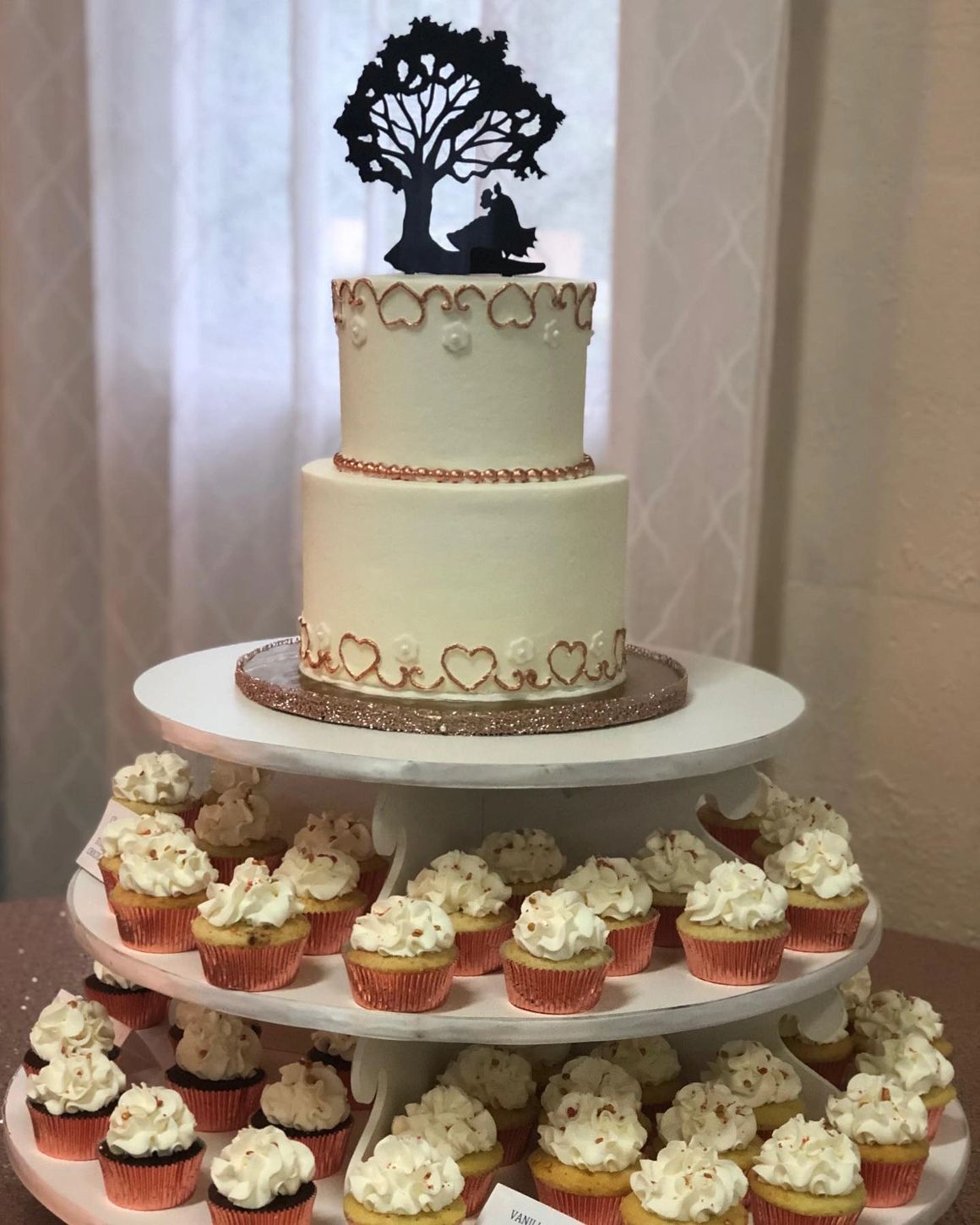 Two-tier white wedding cake with heart decorations and tree silhouette topper, displayed with cupcakes